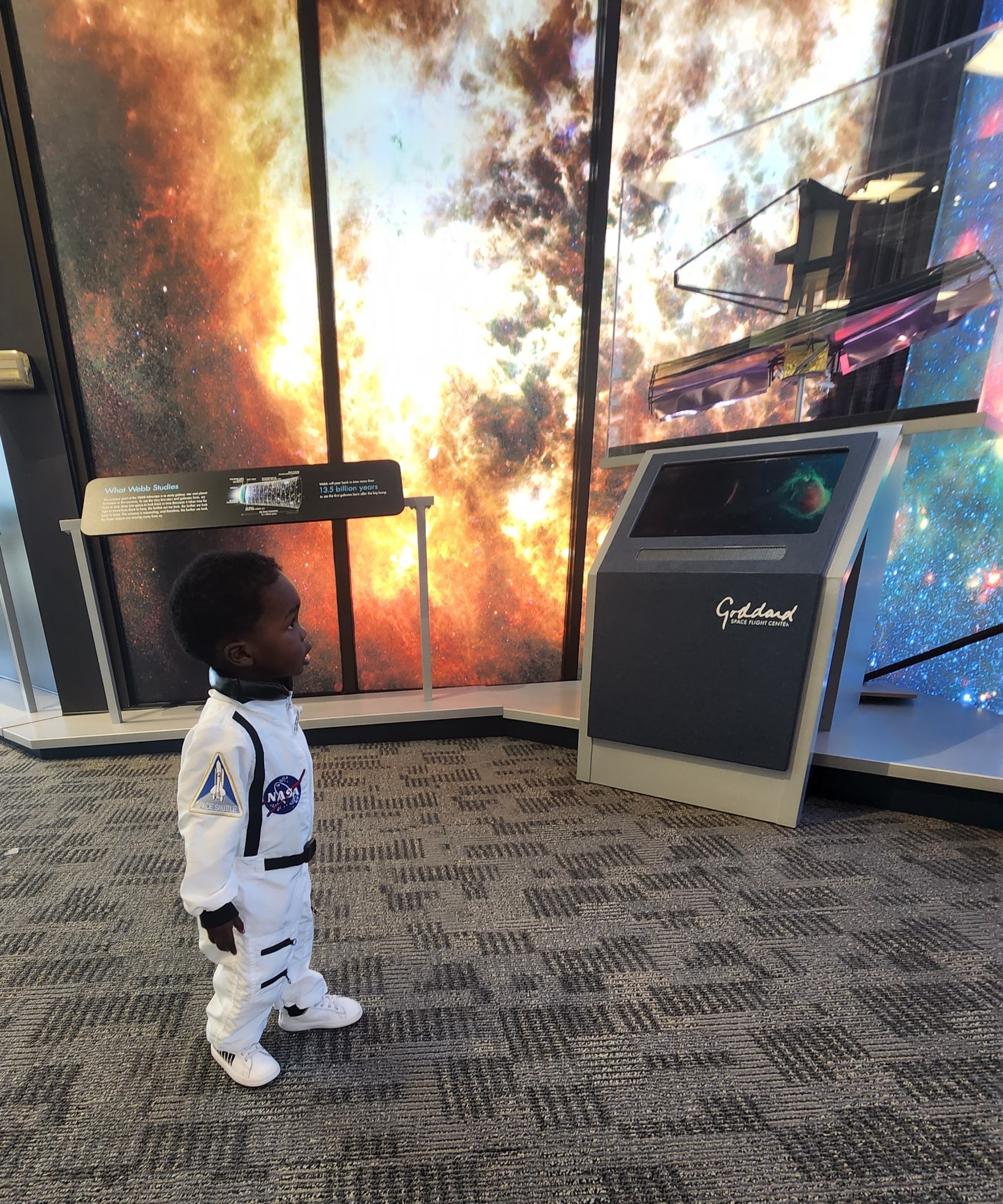 A small boy wearing an astronaut costume stands and looks up at a model of the James Webb Space Telescope. Colorful images of stars and nebulas line the walls behind him.
