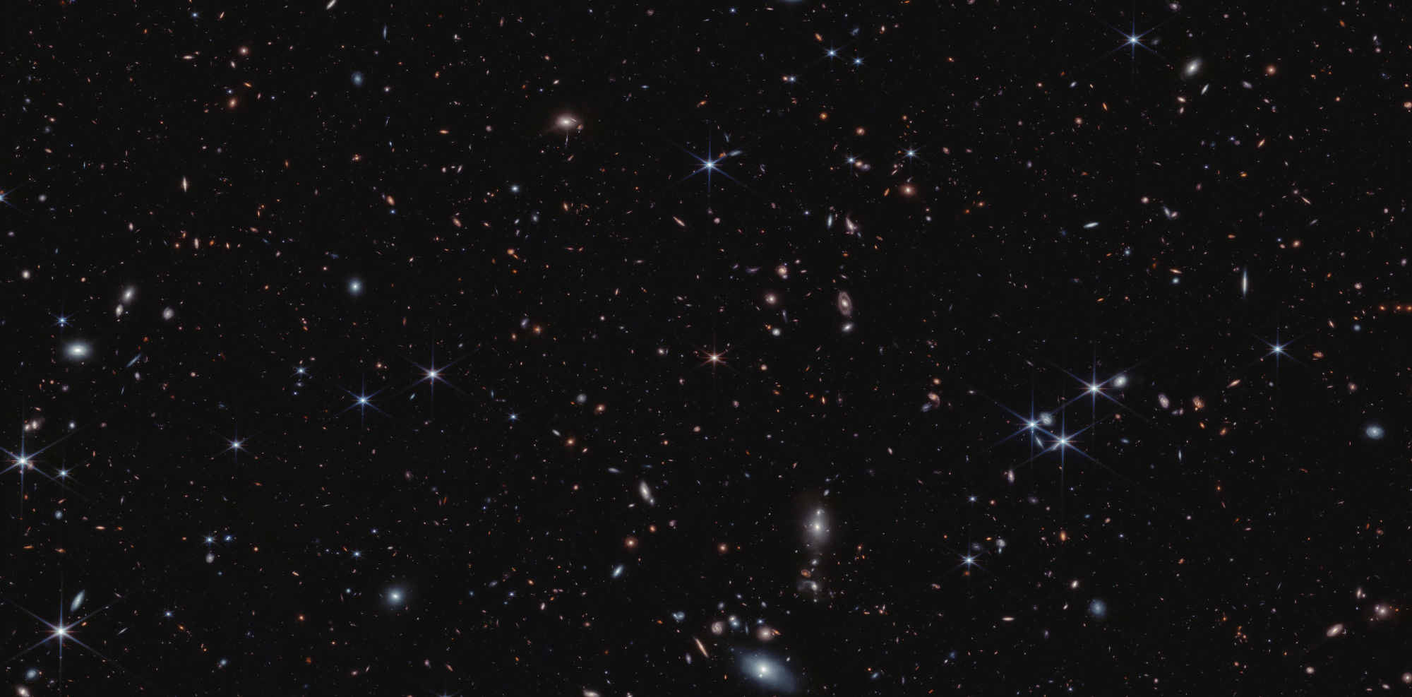 Thousands of tiny colorful galaxies appear across the black expanse of space. At the center is a pink object with six diffraction spikes. It appears slightly smaller than the foreground stars, which appear blue.