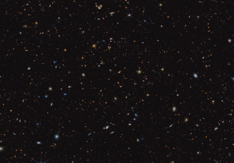 Thousands of small galaxies scattered on a black background. Some are spirals while others are blobby ellipticals. A few spirals are bluish, but most of the galaxies appear yellow or red. A handful of stars display eight-point diffraction spikes.
