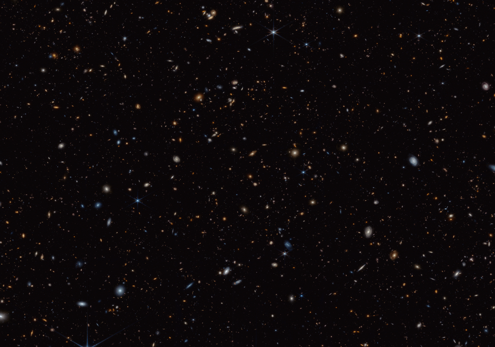 Thousands of small galaxies scattered on a black background. Some are spirals while others are blobby ellipticals. A few spirals are bluish, but most of the galaxies appear yellow or red. A handful of stars display eight-point diffraction spikes.