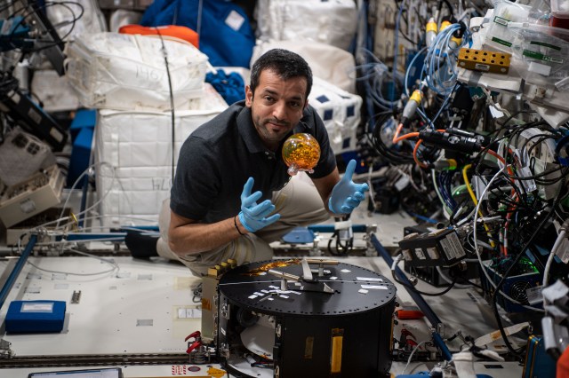 A fist-sized transparent sphere with orange liquid floats out of the hands of Alneyadi, who is facing the camera. He is wearing a black polo shirt. Several cords are against the wall to Alneyadi’s left.