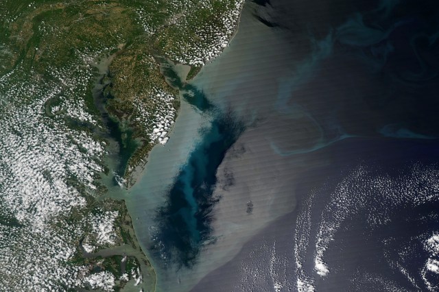 Satellite image of the Delmarva Peninsula area of the East Coast of the U.S., which is a green and brown coastline with the Delaware and Chesapeake Bays. Most of the land is covered in small puffs of clouds. The majority of the image is blue ocean water in the Atlantic. There is a large, gleaming patch of silvery water that looks almost like a mirror, labeled "sunglint." Next to it is an exceptionally colorful patch of ocean water, including teal swirls of phytoplankton, where there is no sunglint, which are labeled "phytoplankton." A darker green and blue patch next to the sunglint is labeled "low wind."