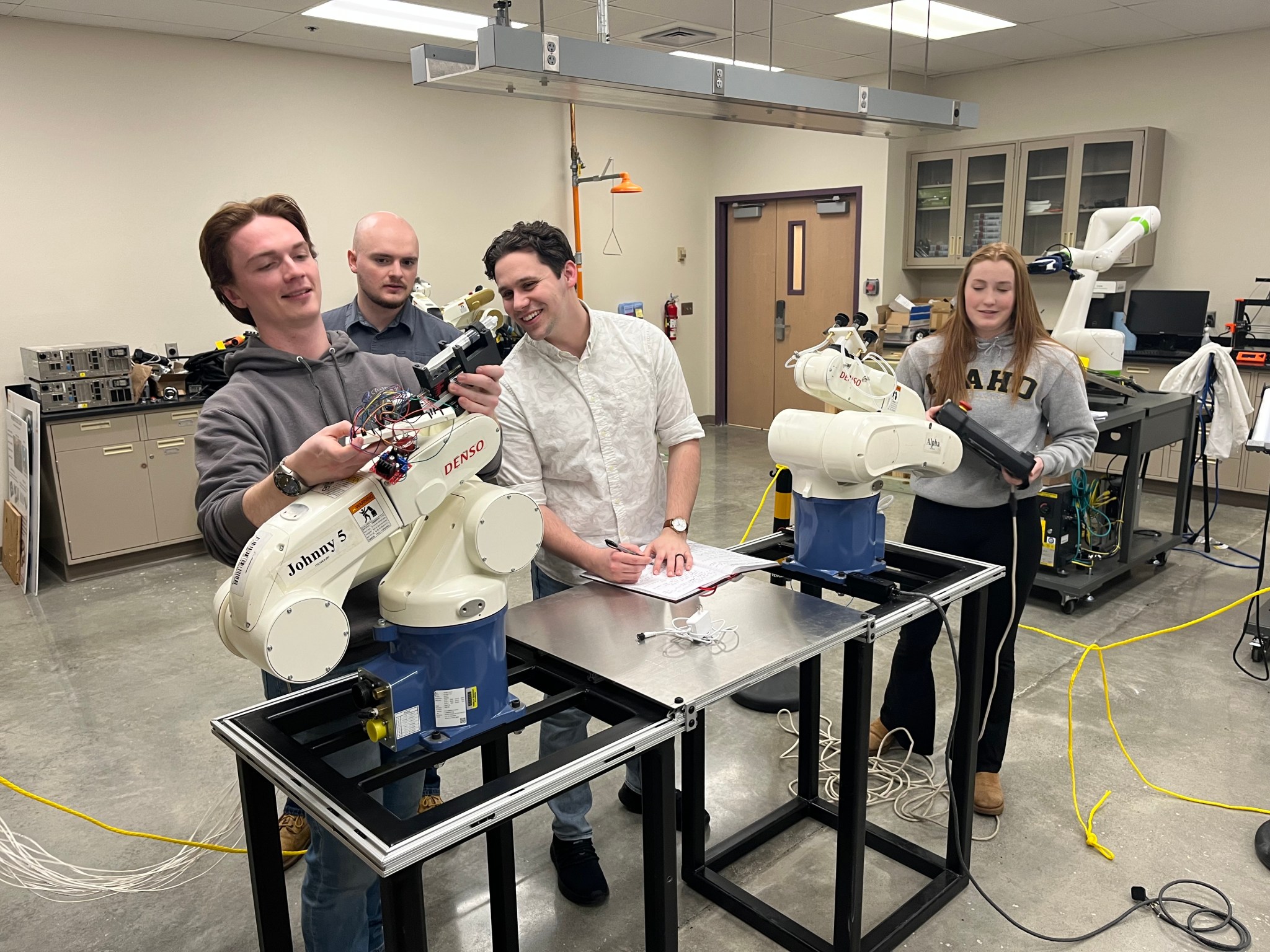 Four students stand around a robotic assembly .