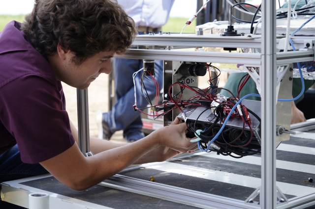 A researcher bends forward to reach into a 3D frame to manipulate the technology suspended in the center via mounting hardware and surrounded by black and red wires and a blue cable.