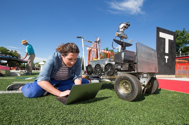 A light-skinned woman with long, dark hair sits on the turf of a football field as she types on a laptop. She is wearing a denim jacket, a white and black striped, blue pants, and black slip-on shoes. Next to her is her Sample Return Robot, which has six wheels, a camera at its front, and a silver metal box as its body. The robot is stationary on a wheeled cart. Behind the woman is a man in a tan, wide-brimmed hat, a light turquoise shirt, and long khaki pants. He is placing an unidentifiable object on a table further in the background. The sky is bright blue with few clouds.
