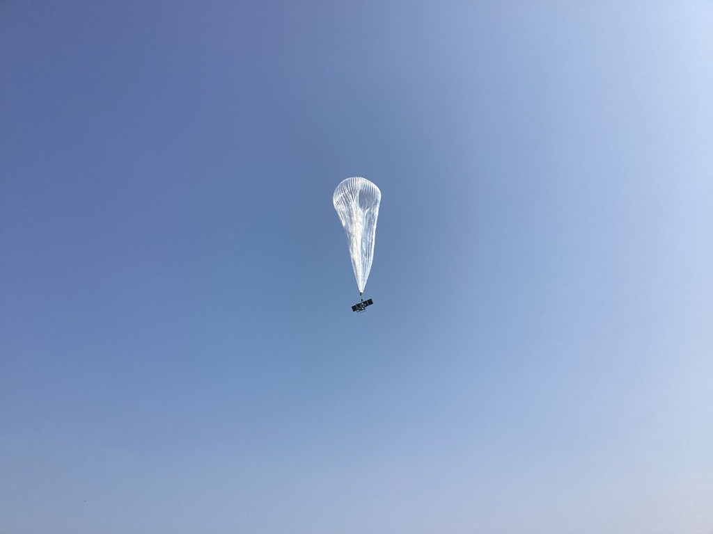 A silvery, translucent balloon with the payload gondola visible beneath it rises into the sky.