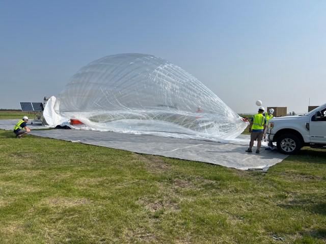 A large translucent balloon resting on a grey tarp in a grassy field swells during inflation. Crew members wear yellow safety vests and white hard hats near the front end of a white truck.