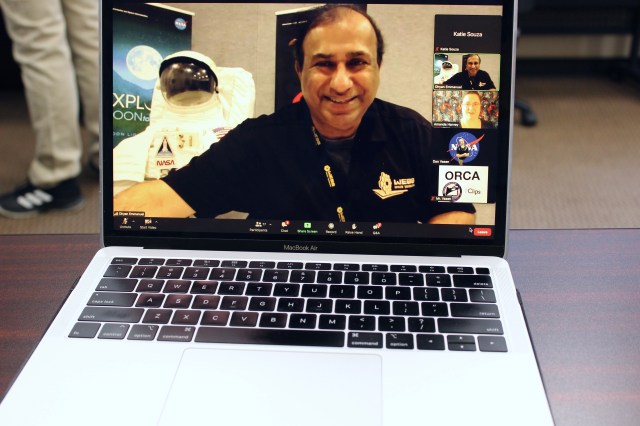 A silver laptop sits on a table with a view of a Zoom-like call. A person takes up the larger view in the call, with four other smaller views on the right side.