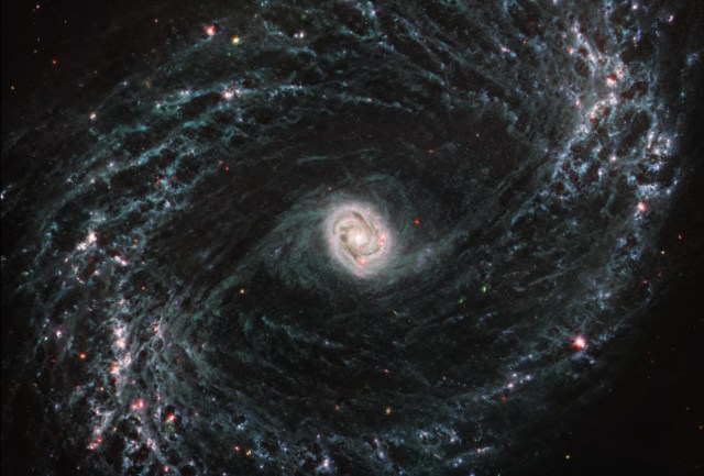 This image taken by the NASA/ESA/CSA James Webb Space Telescope shows one of a total of 19 galaxies targeted for study by the Physics at High Angular resolution in Nearby Galaxies (PHANGS) collaboration. Nearby barred spiral galaxy NGC 1433 takes on a completely new look when observed by Webb’s Mid-Infrared Instrument (MIRI). NGC 1433’s spiral arms are littered with evidence of extremely young stars releasing energy and, in some cases, blowing out the gas and dust of the interstellar medium they plough into. Areas that once appeared dark and dim in optical imaging light up under Webb’s infrared eye, as clumps of dust and gas in the interstellar medium absorb the light from forming stars and emit it back out in the infrared. Webb’s image of NGC1433 is a dramatic display of the role that dynamic processes within the forming stars, dust, and gas play in the larger structure of an entire galaxy. At the centre of the galaxy, a tight, bright core featuring a unique double ring structure shines in exquisite detail, revealed by Webb’s extreme resolution. In this case, that double ring is actually tightly wrapped spiral arms that wind into an oval shape along the galaxy’s bar axis. NGC 1433 is classified as a Seyfert galaxy, a galaxy relatively close to Earth that has a bright, active core. The brightness and lack of dust in the MIRI image of NGC 1433 could hint at a recent merger or even collision with another galaxy.In the image of NGC 1433, blue, green, and red were assigned to Webb’s MIRI data at 7.7, 10 and 11.3, and 21 microns (the F770W, F1000W and F1130W, and F2100W filters, respectively). MIRI was contributed by ESA and NASA, with the instrument designed and built by a consortium of nationally funded European Institutes (The MIRI European Consortium) and NASA’s Jet Propulsion Laboratory, in partnership with the University of Arizona.[Image Description: A large galaxy takes up the entirety of the image. The core is mostly bright white, but there are also swirling, detailed structures that resemble water circling a drain. There is white and light blue colored dust that emanates from the core’s center, but it is tightly contained to the core. The rings are wispy and highlight filaments of dust around cavernous black bubbles. The dust in the outer rings contains dots that are navy blue, pinkish, reddish, and white.]