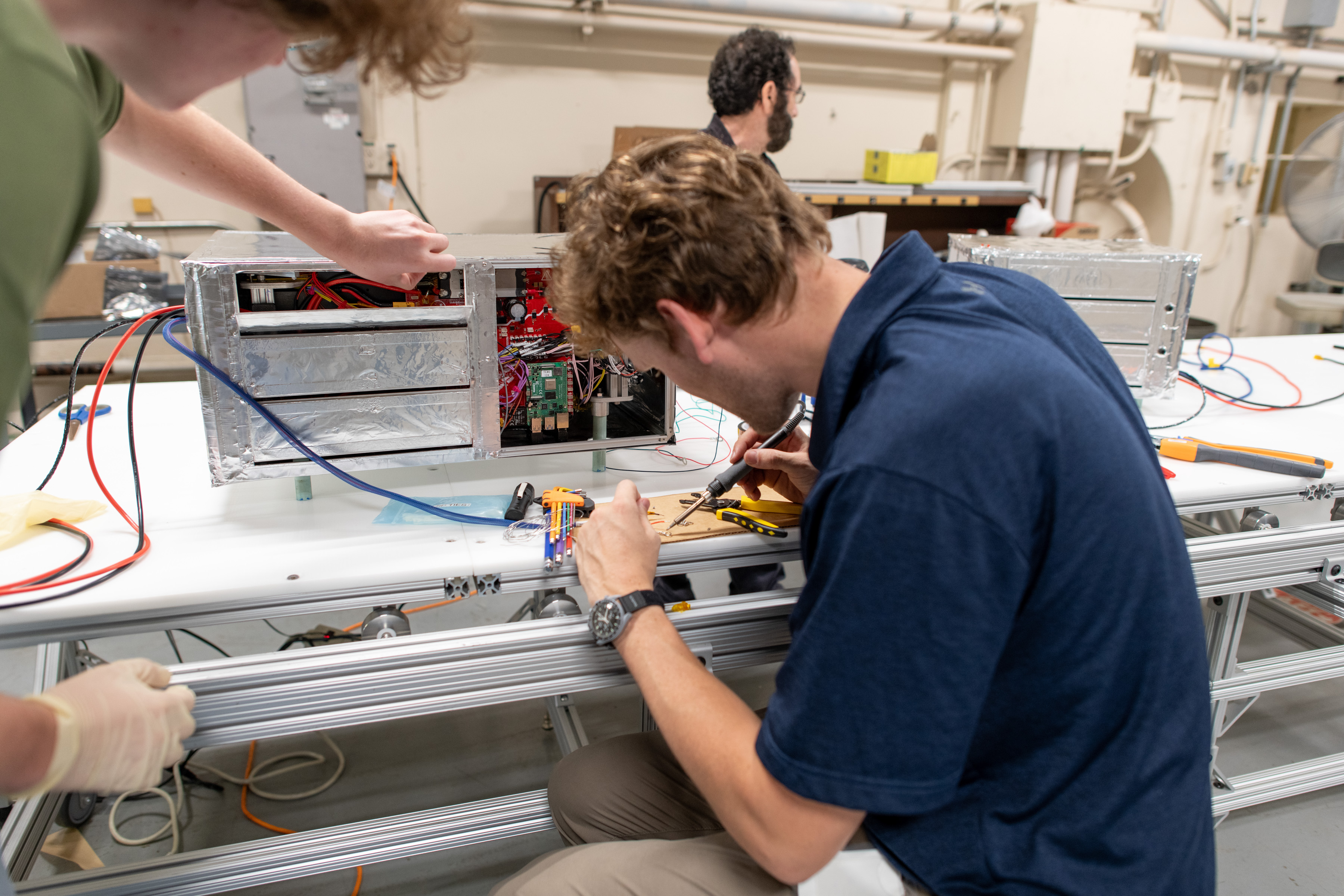 A man at the forefront of the photo sits with his back turned to the viewer, using a soldering iron as he works on a project. On the table is hardware in the form of a box covered in silver-colored foil.