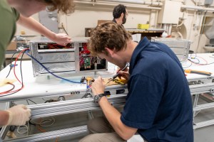 A man at the forefront of the photo sits with his back turned to the viewer, using a soldering iron as he works on a project. On the table is hardware in the form of a box covered in silver-colored foil.