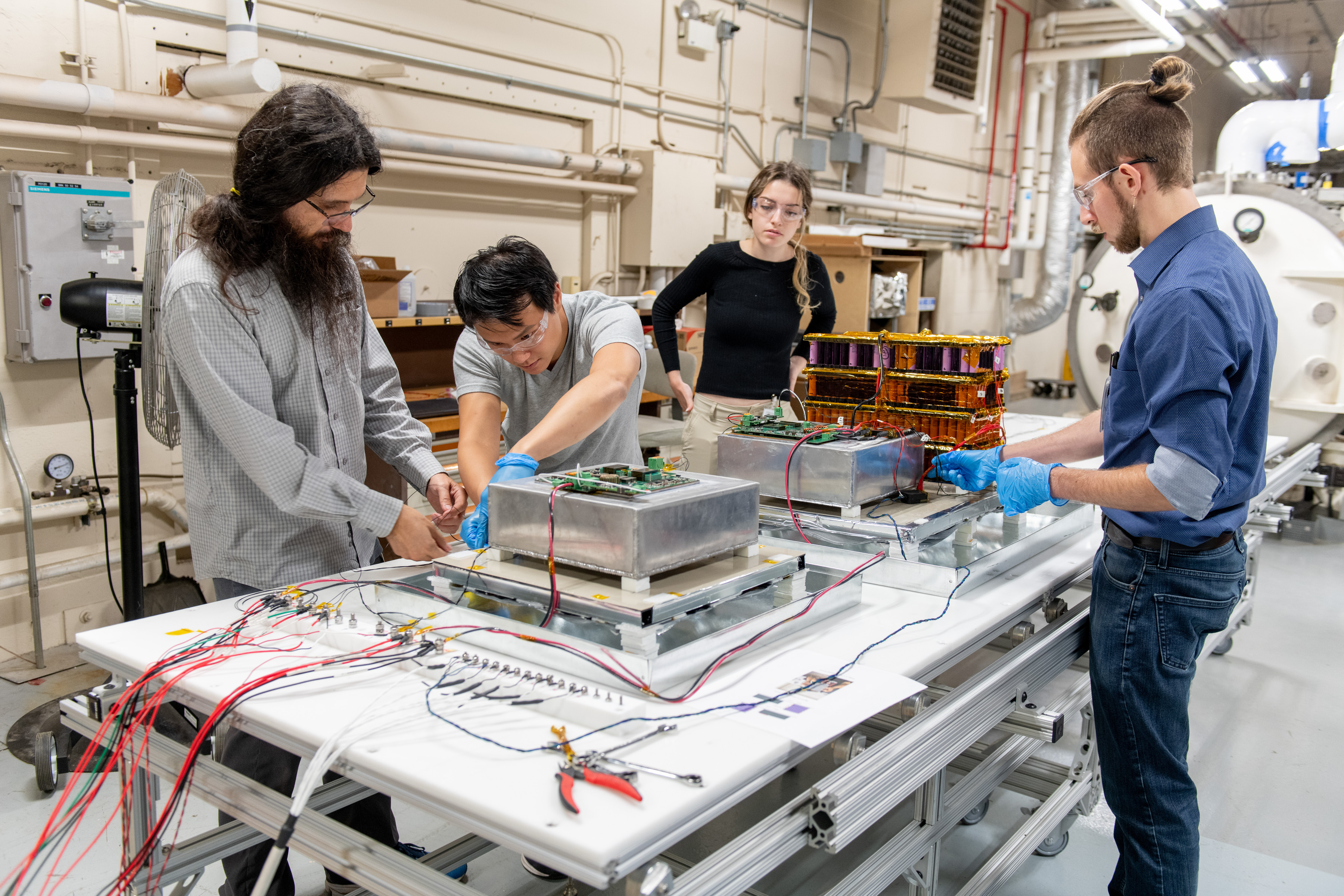 Four people stand around a table working on various parts of a hardware prototype that includes wires, system boards, and silver-colored boxes. In the background is NASA's vacuum chamber, which was used to test the hardware.