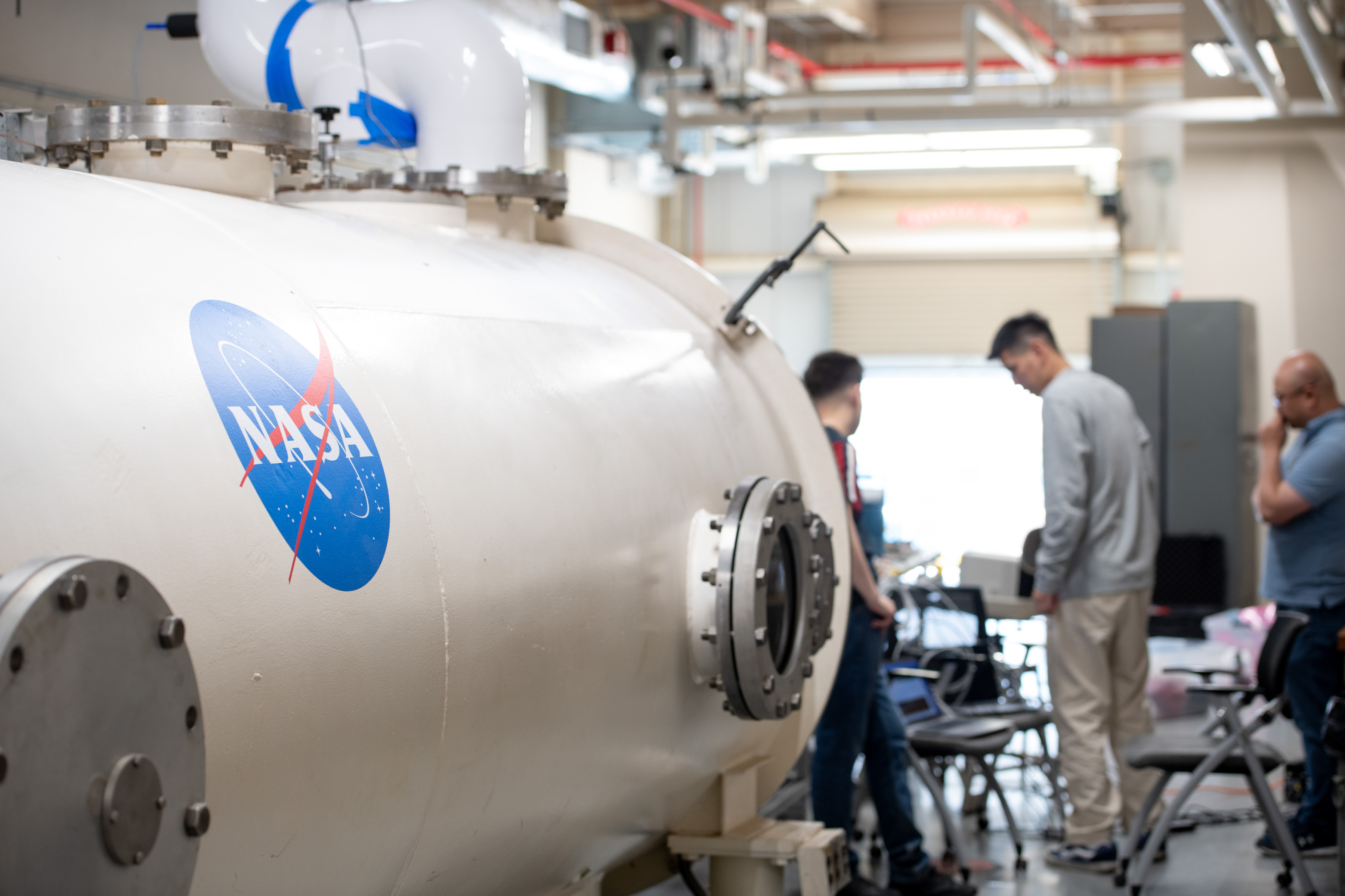 In the foreground taking up the left half of the photo is a cylindrical vacuum chamber with the NASA insignia printed on the side. Blurred in the background are people looking at a laptop and other technology obscured from view.