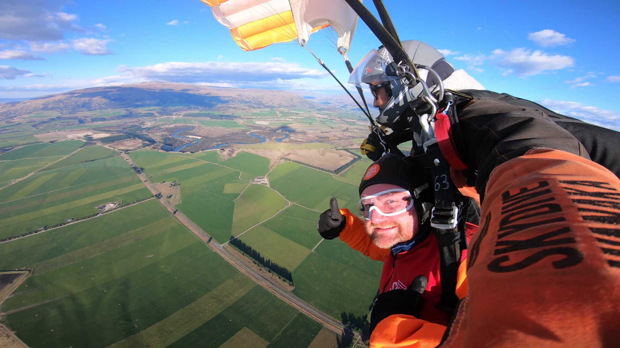 A wide-angle image of a man skydiving over green fields and brown hills, with bright blue sky above him and white clouds below him. The camera is mounted on the skydiving rig and the man is distorted. He wears orange and a black hat.
