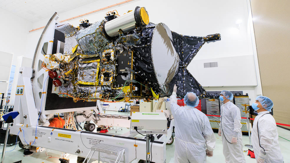 Three people inspect a partially assembled spacecraft.