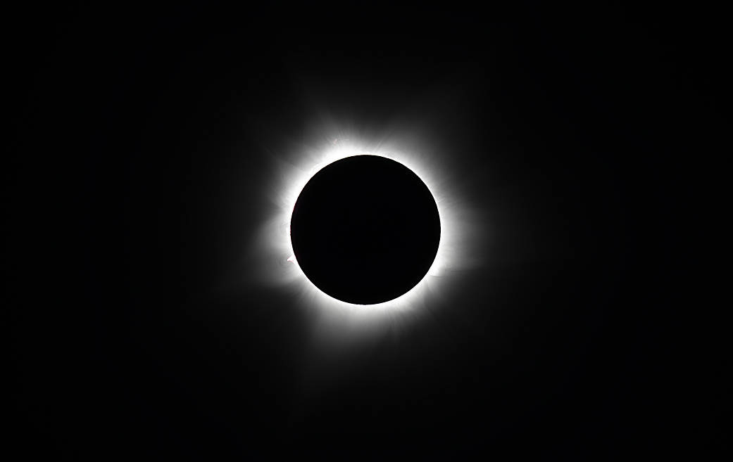 Against a black background, a black circle (the Moon) is surrounded by a streams of white light (the Sun's corona). In the bottom left corner, a small peak is visible coming off of the circle – a solar prominence.