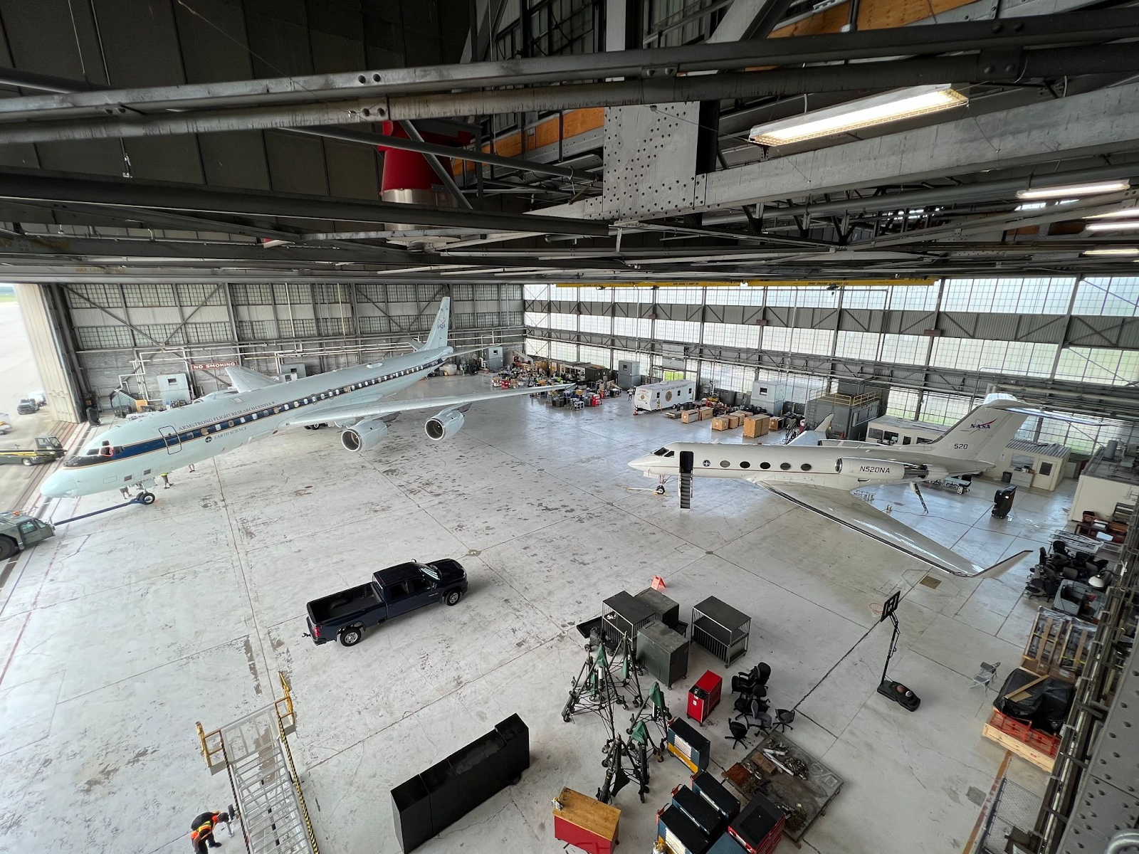 Two research planes are parked in a spacious aircraft hangar lined with cargo. Several people, a black pickup truck, and even a basketball hoop – for recreation during down time – are also visible.