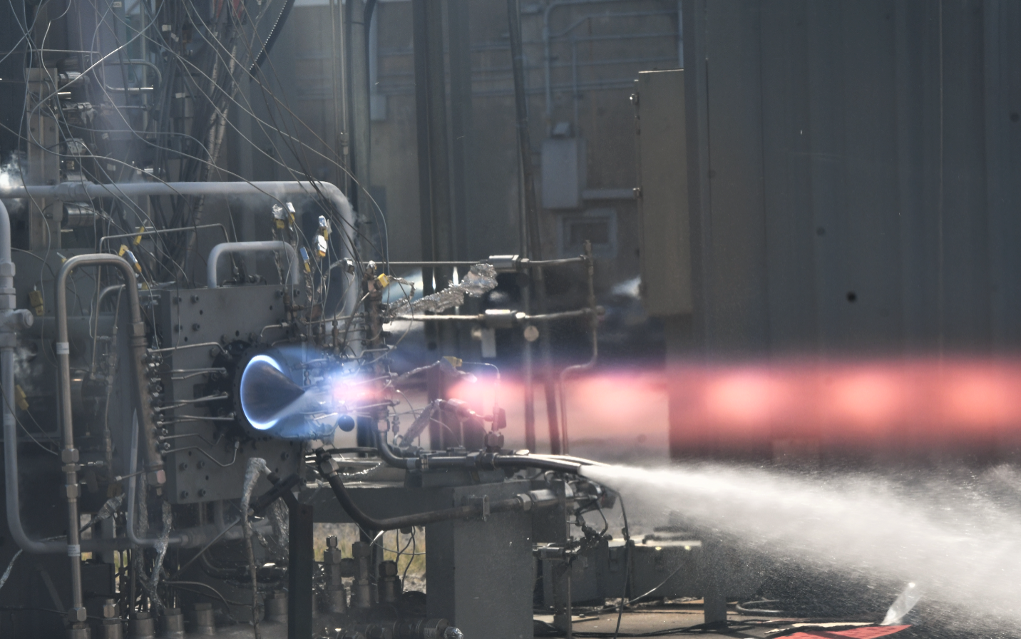 Rotating detonation rocket engine, or RDRE hot fire test at Marshall Space Flight Center. On a metal test stand, the engine appears to have a ring of blue light at the base, and the flames turn redish orange as they are ejected from the rocket engine.
