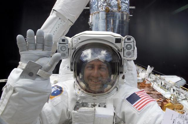 An Astronaut in a spacesuit smiles while waving at the camera while on a spacewalk.