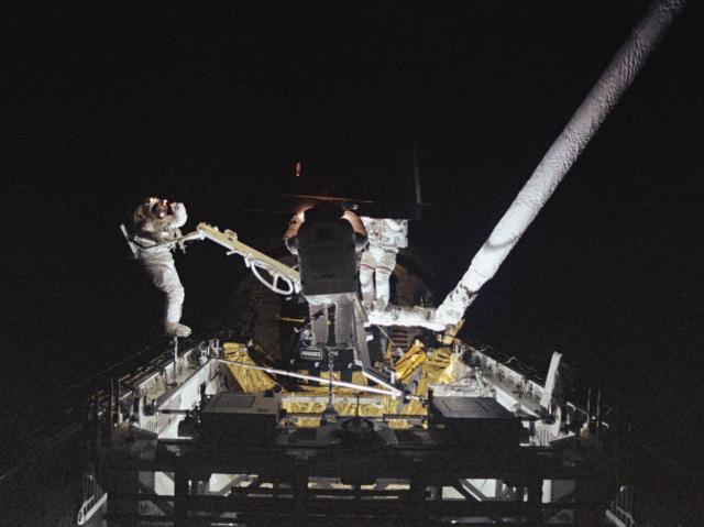 Three Astronauts on a spacewalk in the dark work to pull a satellite into the space shuttle.