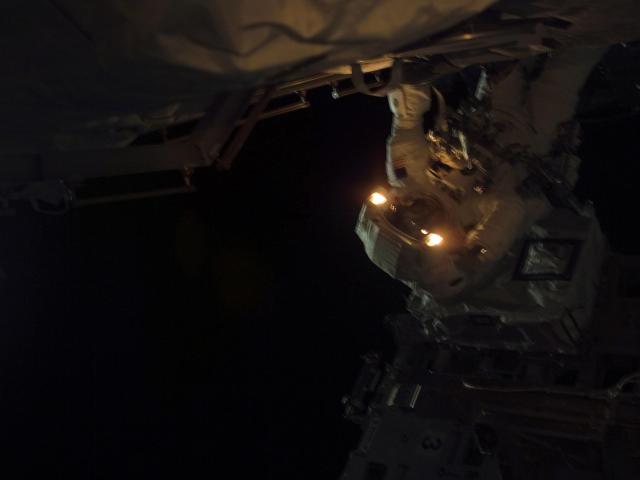 An astronaut works in the dark, with the lights in their suit on.