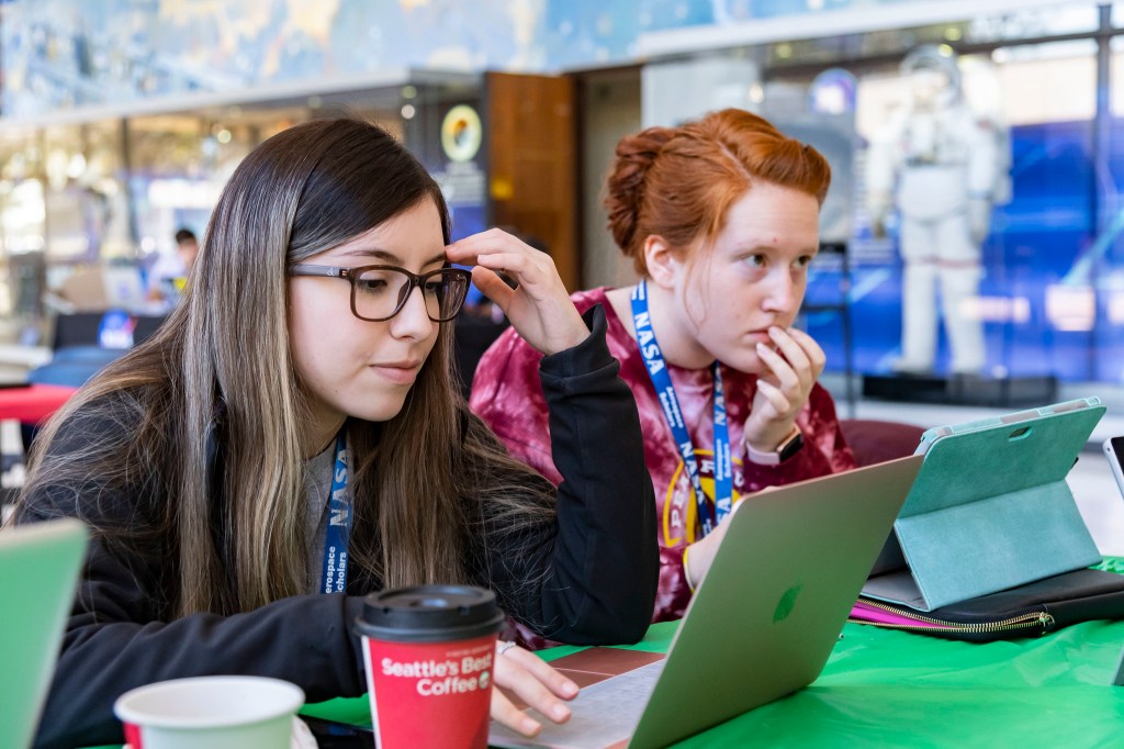 Two NASA Community College Aerospace Scholars sit at a desk and work on projects on laptops sitting in front of them. The duo wears casual attire and have NASA lanyards on. In the background a display of a NASA spacesuit is out of focus. Credit: NASA/Robert Markowitz