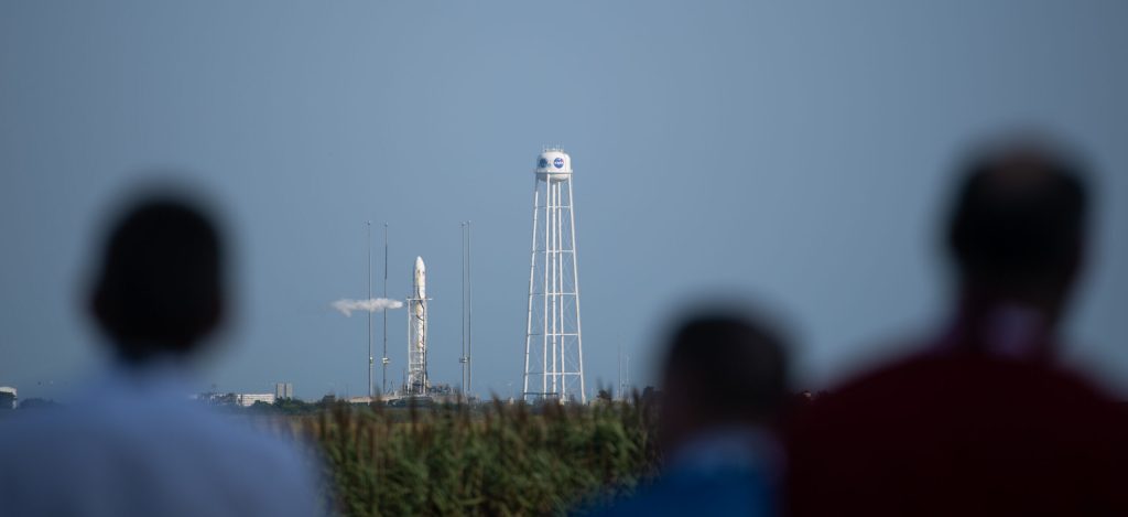 A large, white rocket stands vertical on a launch pad, a thin trail of white exhaust exiting toward the middle of the rocket. A tall, white water tower stands next to the launch pad against a pale blue sky. In the foreground, blurred out, are the heads of shoulders of people looking at the rocket.