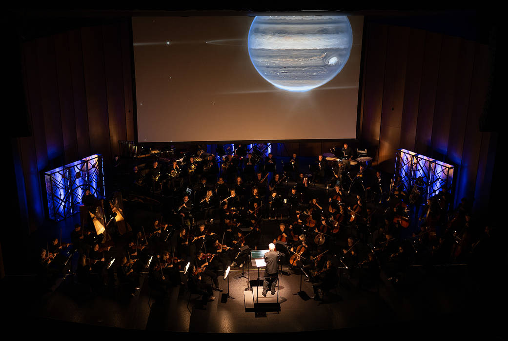 An image of Jupiter is projected large on a screen above an orchestra on a darkened theater stage. The musicians are barely visible, with a soft light on the conductor. Blue lights illuminate the back of the stage.