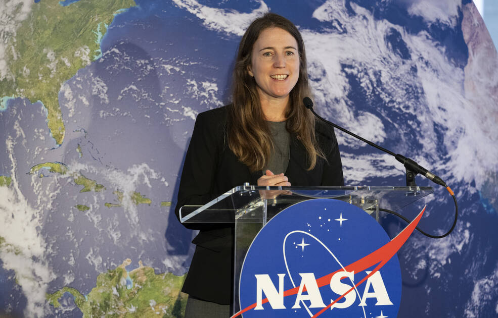Kate Calvin stands behind a clear podium with the NASA meatball logo. She is addressing a crowd. She is framed in front of a satellite image of Earth.