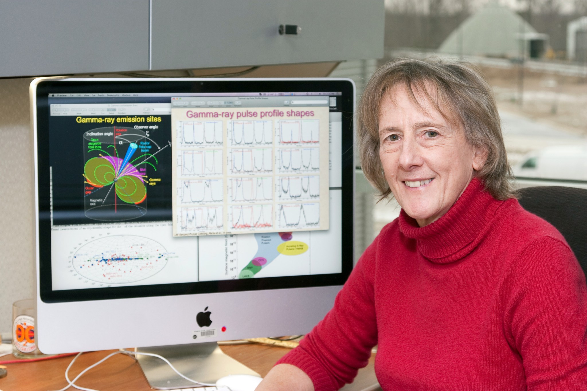 At left is a computer screen with information about pulsars. At right sits a a woman in a red pullover top facing the camera.