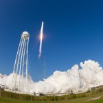 A large, white rocket lifts off from a launch pad against a bright blue sky. White flame and exhaust streams out from its base with billowing clouds of exhaust on the launch pad. A tall water tower stands next to the launch pad. In the foreground, tall green grasses behind a fence line.