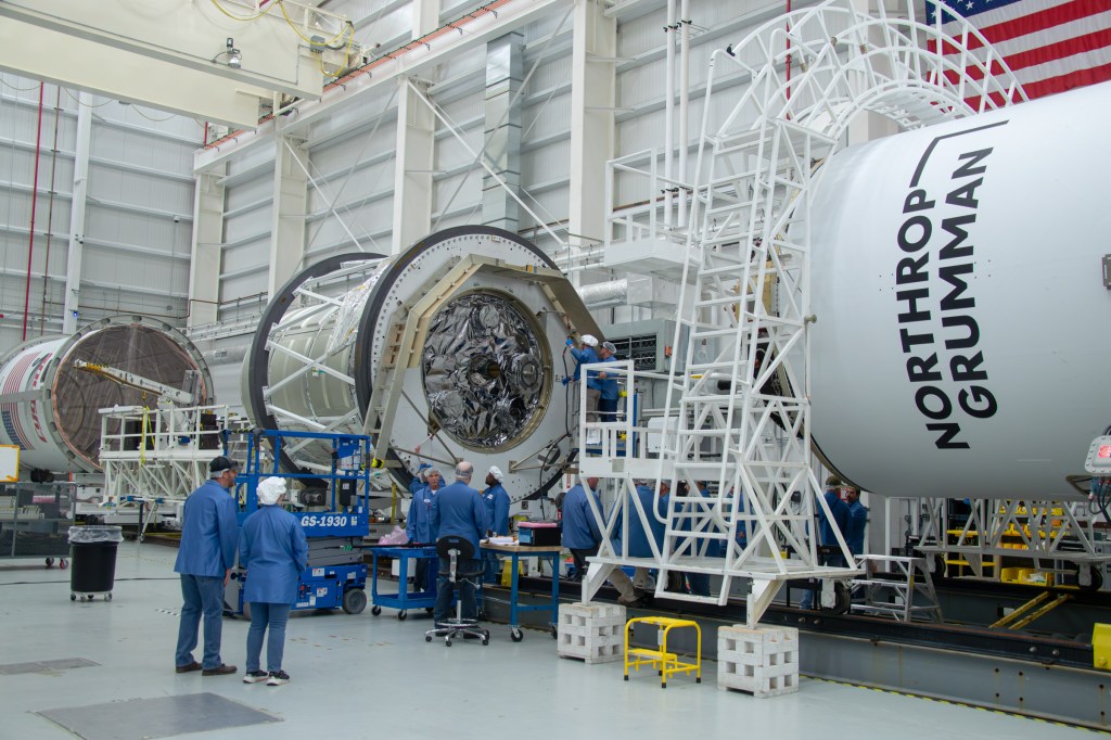 The Cygnus spacecraft and Antares rocket in a large white warehouse room. On the far left, the top part of the Antares rocket that will cover the Cygnus spacecraft is a white empty cylinder. Closer to the center, another component of the spacecraft is a silver, foil-wrapped cylinder with silver hoops around it. On the right, the top of the white Antares rocket is visible, with Northrup Grumman posted on the side. People in blue lab coats and white hair nets are scattered around the room, examining pieces of the spacecraft.