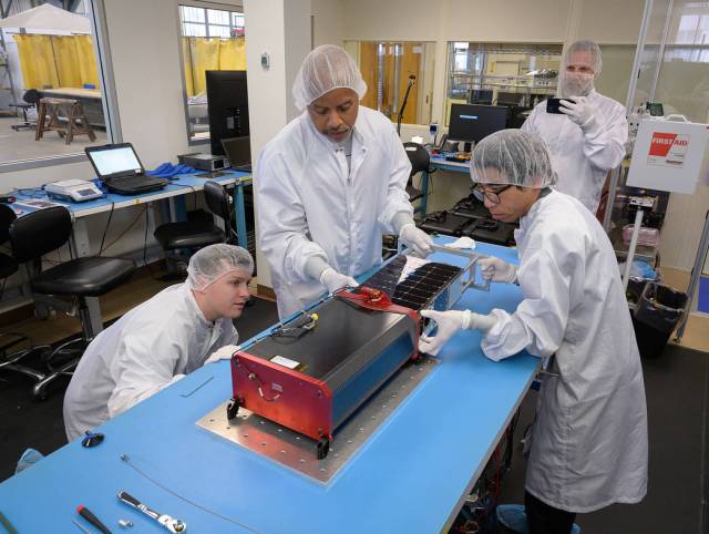 Two team members load a spacecraft into a CubeSat dispenser in a cleanroom. Two other team members observe.
