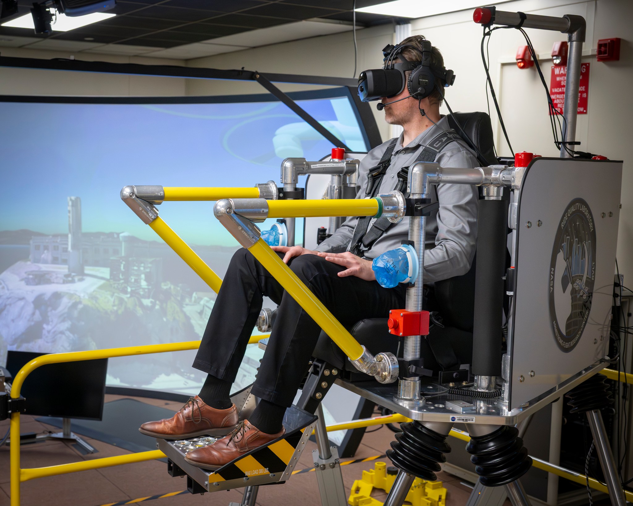 A man sits in a seat with straps atop a metal platform. His feet rest on a metal platform. Yellow metal bars surround the platform. The man is wearing a gray shirt, black pants, and brown shoes, as well as black virtual reality goggles and a headset. To the left of the man is a large screen that shows the former prison structure of Alcatraz Island in San Francisco Bay.