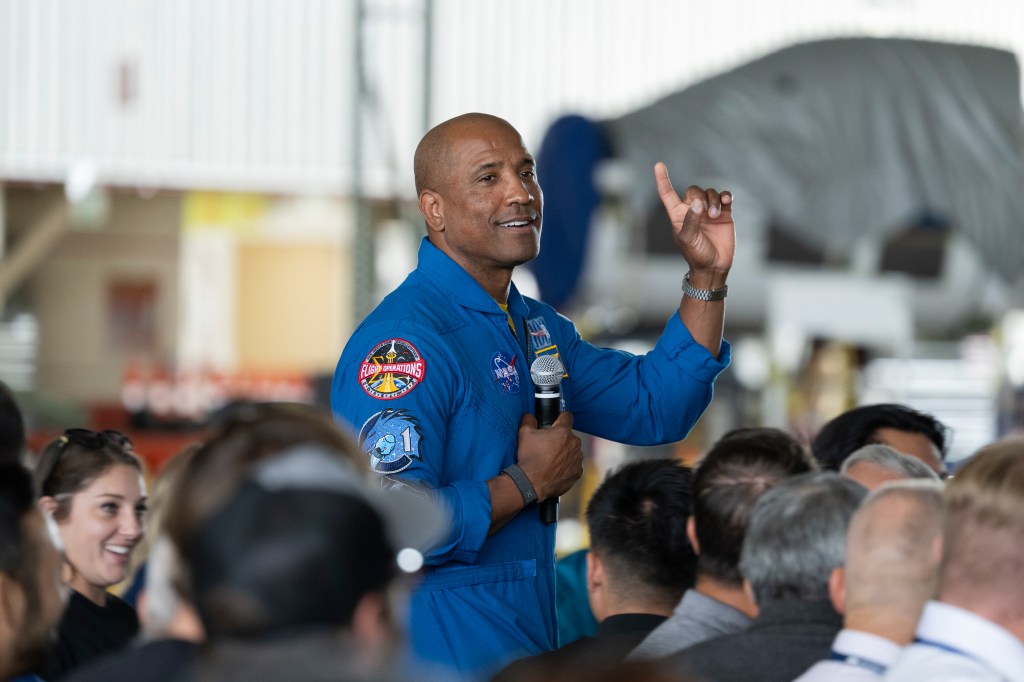 A NASA astronaut wearing a bright blue flight suit speaks to a large audience inside an aircraft hangar.