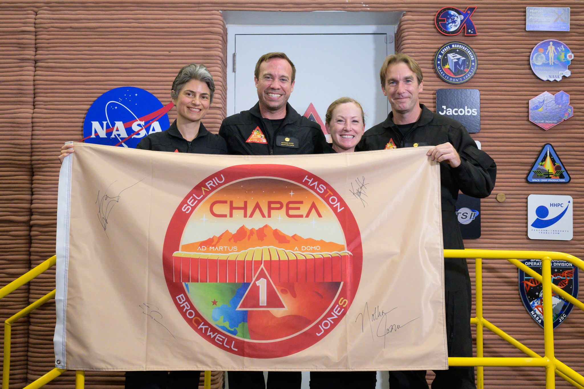 The CHAPEA mission 1 crew poses with a flag featuring their mission patch surrounded by their signatures.