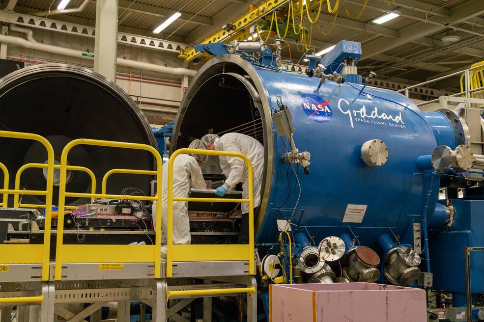 People in white clean suits and hair nets stand on a yellow platform with railings in front of a large blue cylinder.