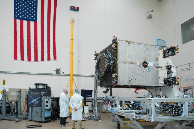 Two scientists in lab coats stand with the OSAM 1 robot with an American Flag on the wall to the left