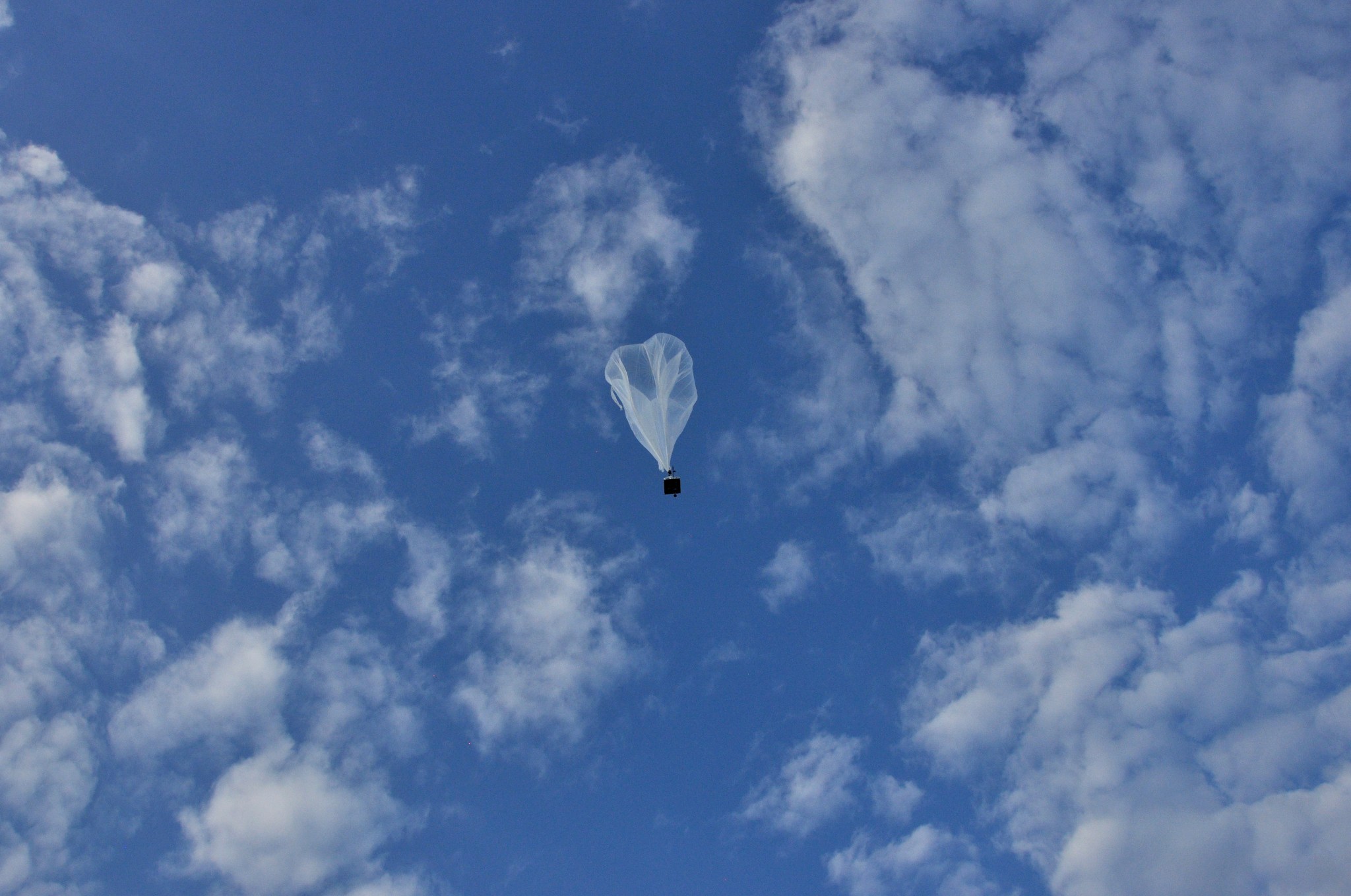 White/translucent balloon carrying a small rectangular load in center frame makes its ascent. Blue sky dotted with light clouds in background.
