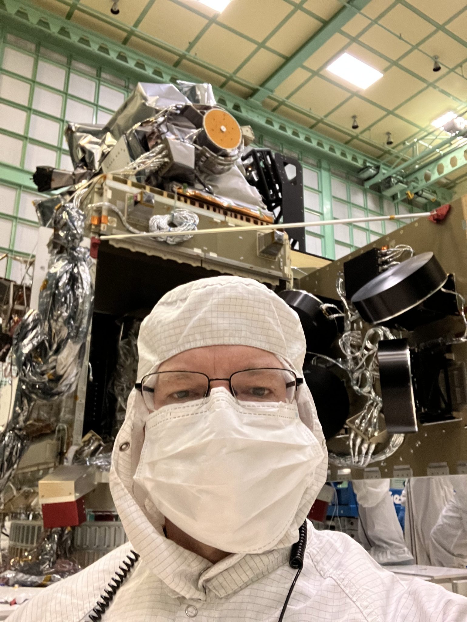 A man with glasses, wearing white hooded coveralls and a white mask, takes a selfie in front of a spacecraft in a clean room. The spacecraft is gold and sits on a tall table. The walls are gridded with filter panels.