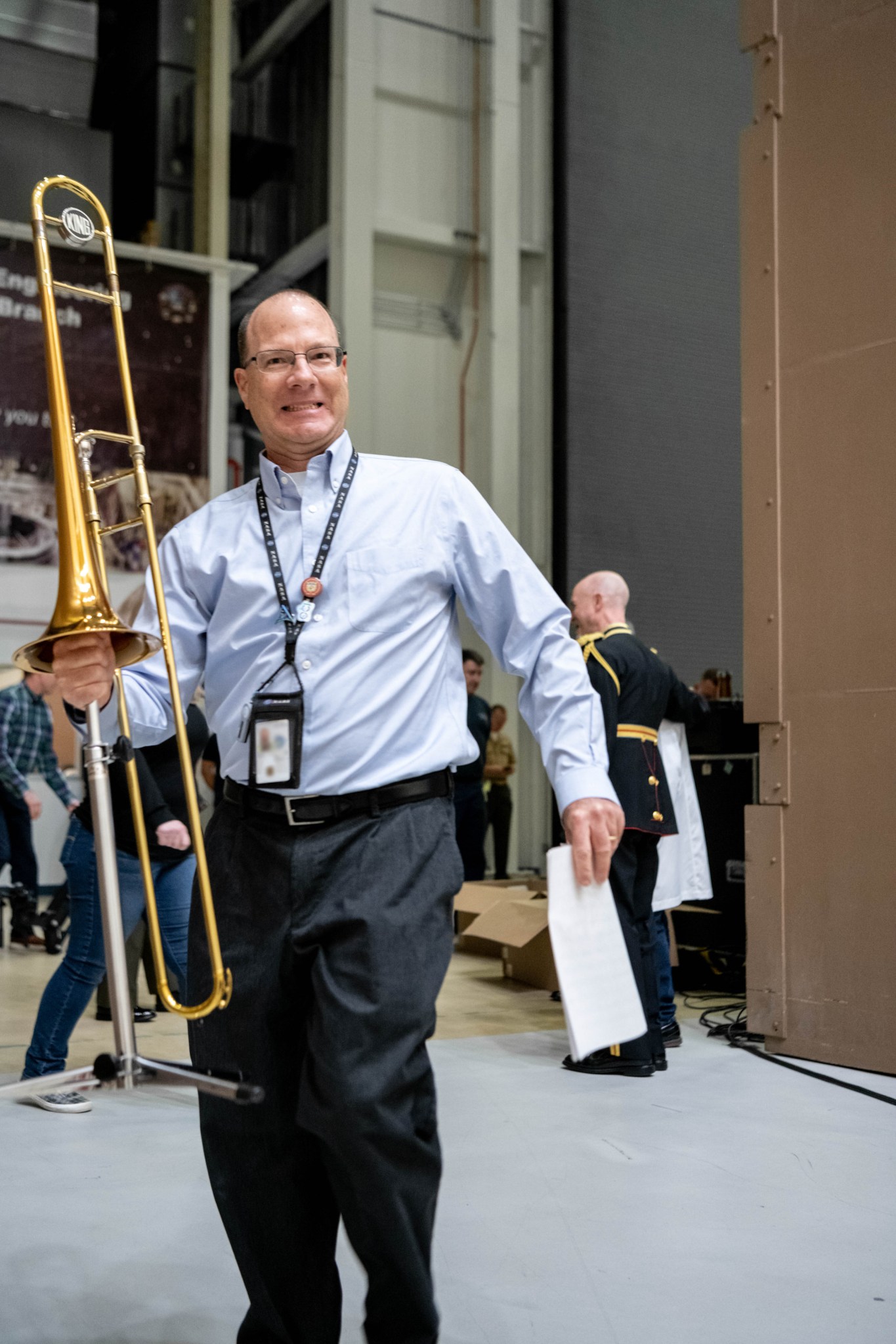 Gary Davis, a man in a blue shirt, dark pants, glasses, and a lanyard, smiles broadly while holding a trombone in Goddard's acoustic testing chamber. Other musicians are visible behind him.