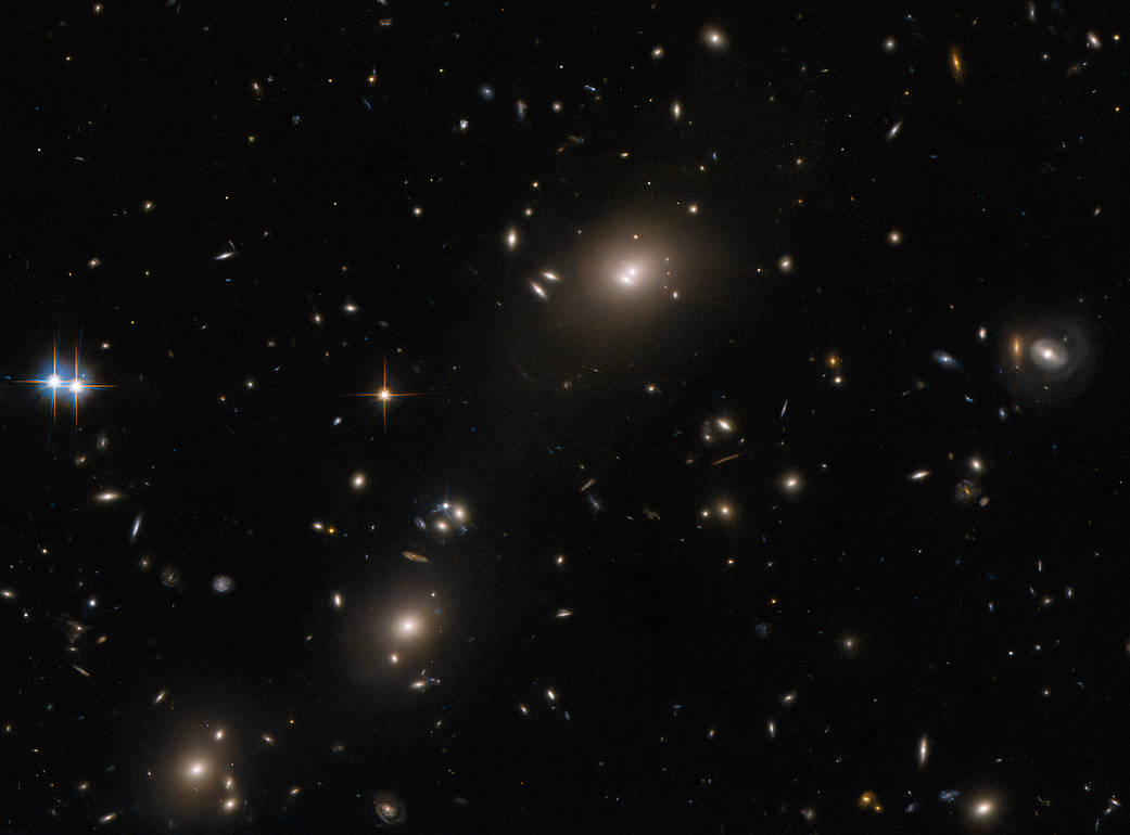 Oval-shaped, elliptical galaxies. Largest has 2 bright spots in its core. It and 2 others look like galaxy clusters, surrounded by smaller galaxies. Left edge: two bright stars with four long spikes. Right edge: small ring-shaped galaxy.