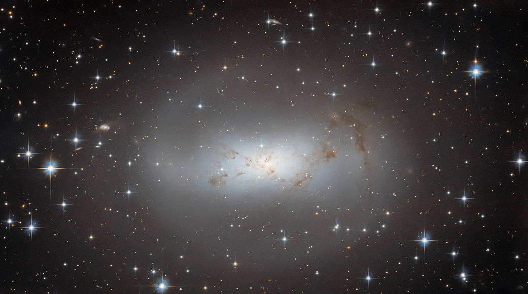 A large, bright, diffuse galaxy. Center is brighter and bluer, fading to a pale, faint, gray halo. An arm on one side that curls around the top. Threads of dark dust cross the center. Many stars shine around the galaxy, on a black background.