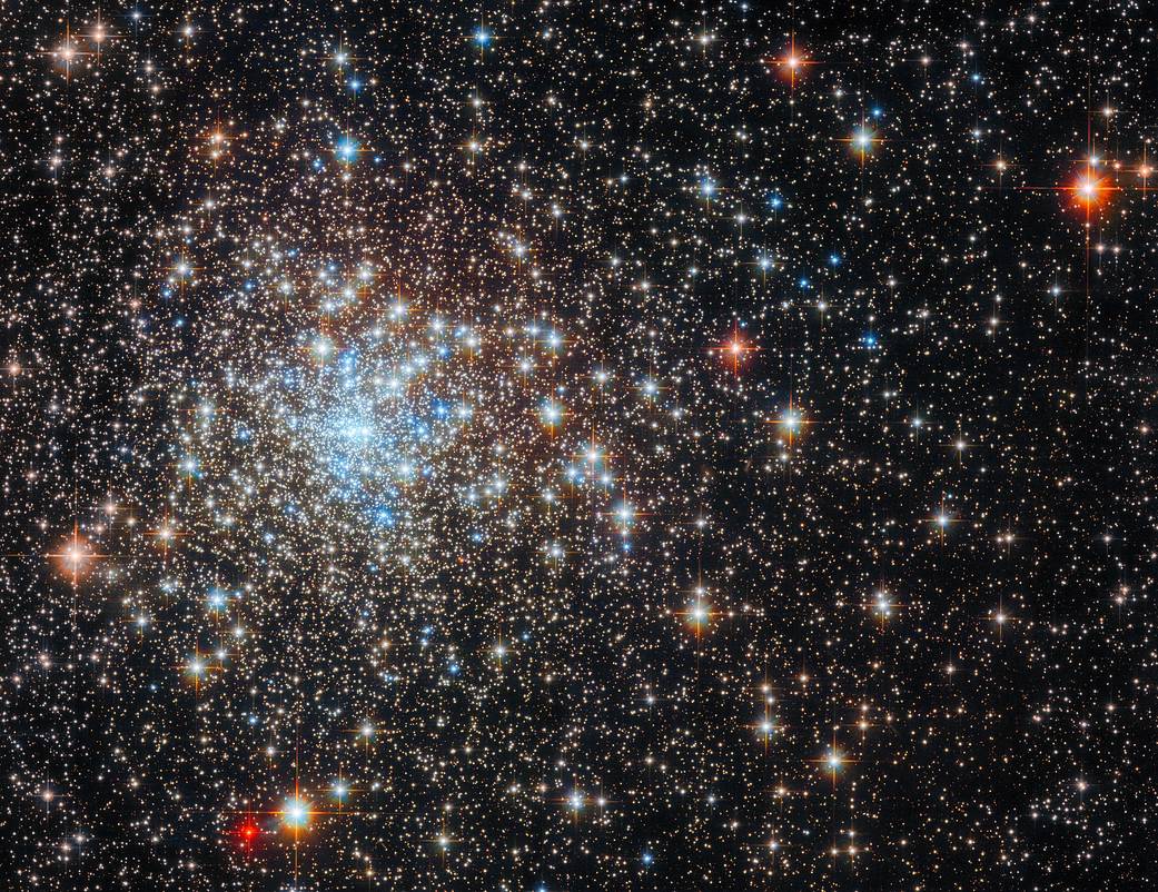 Dense cluster of bright stars. Its core is to the left and has a distinct group of blue stars. Surrounding the core are stars in warmer colors. They are numerous near the core, becoming more sparse, small, and distant toward the sides of the image.