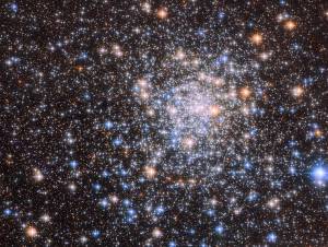 The view is filled with small stars, on a dark background. Stars become much denser and brighter around a core just right of center. Most of the stars are small, but some are larger with a round, brightly colored glow and four sharp diffraction spikes.