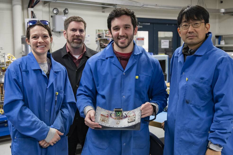 four individuals stand in a lab wearing blue and black lab coats and smiling.