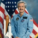 An astronaut posing in front of the American flag with a model of the Space Shuttle.