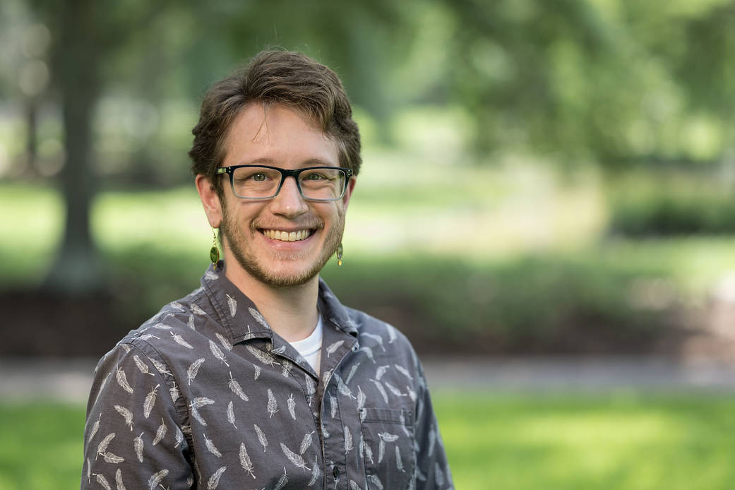 Mars Research Scientist S. J. Ralston smiles at the camera. They are wearing glasses, earrings, and a collared shirt. The background is out of focus.