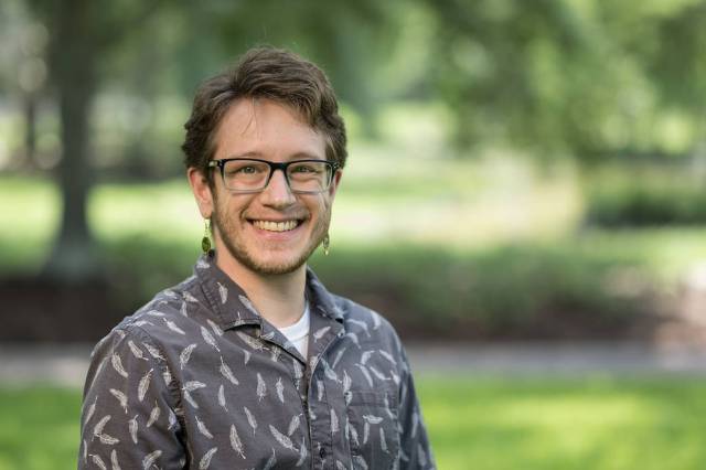 Mars Research Scientist S. J. Ralston smiles at the camera. They are wearing glasses, earrings, and a collared shirt. The background is out of focus.