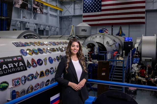 A woman with long, curly, and brown balayaged hair smiles widely, wearing a long black suit jacket and white shirt. She's standing in front of space hardware with International Space Station mission patches on showcase and an American flag behind her.
